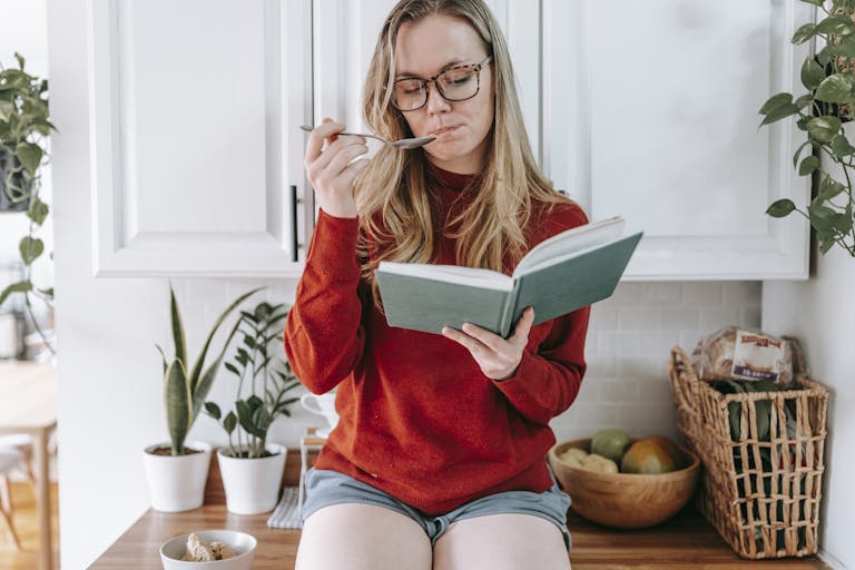 A Woman Eating while Reading a vegan Book in the Kitchen