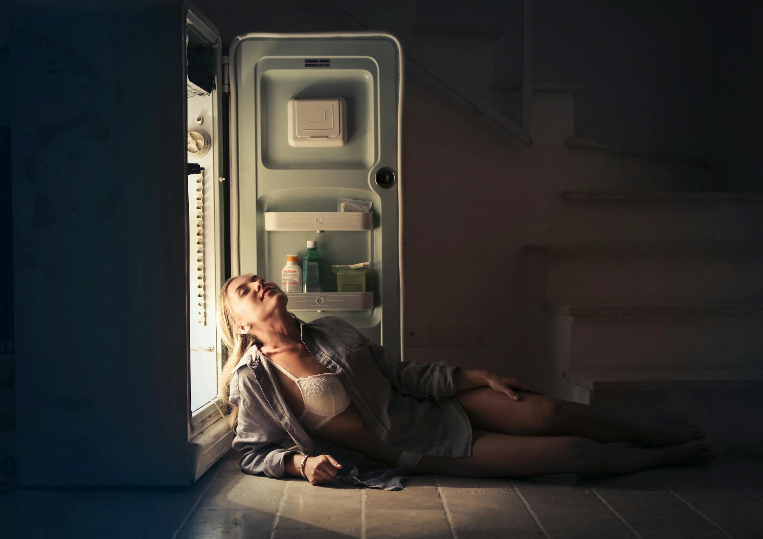 A woman relaxes near an open refrigerator in a dimly lit room, creating a serene and moody atmosphere.