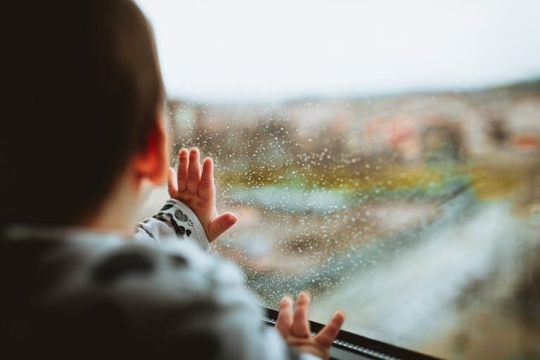 A young child gazes outside through a rainy window, capturing a moment of curiosity and wonder.
