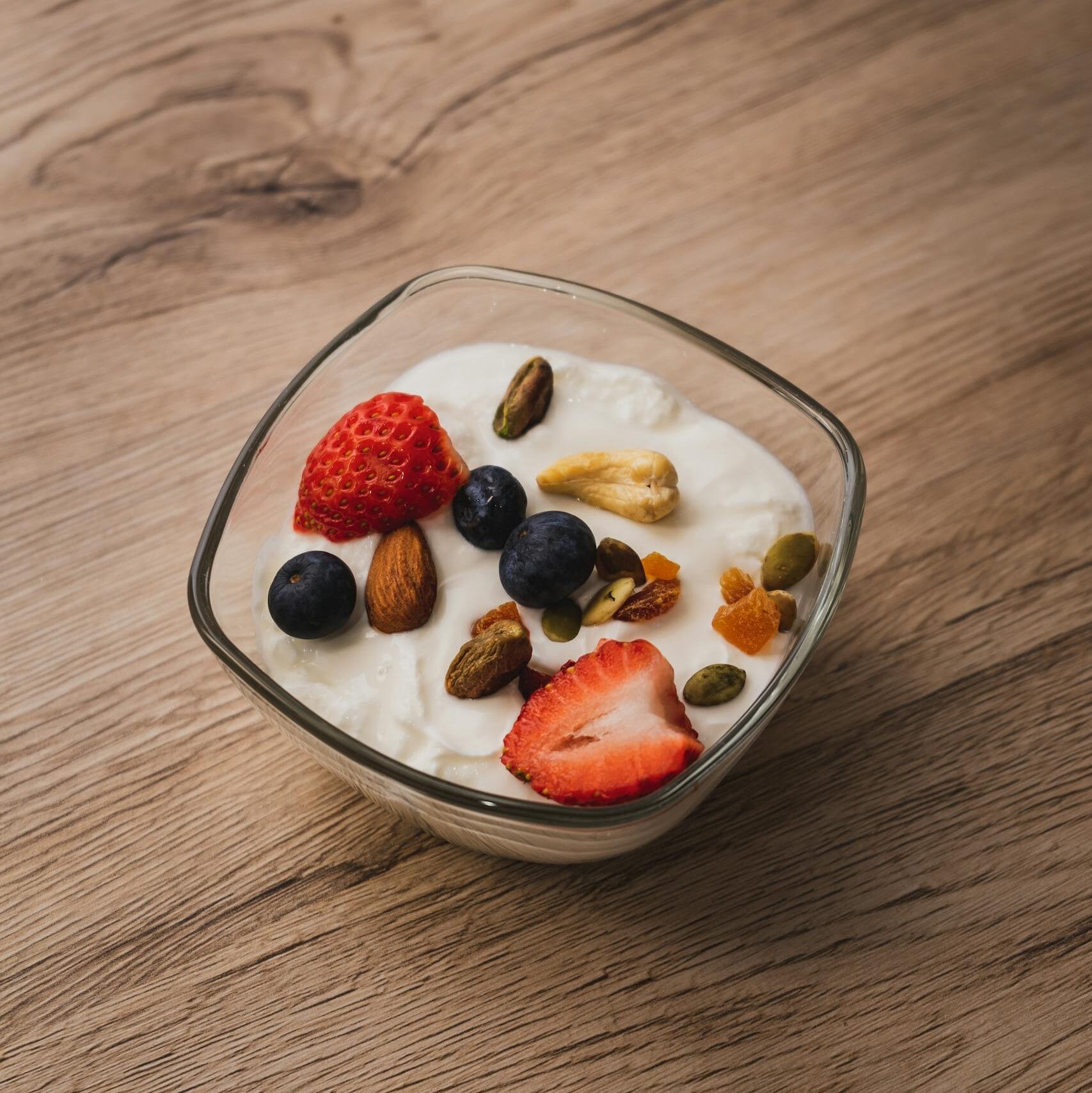 A bowl of yogurt topped with fresh strawberries, blueberries, and assorted nuts on a wooden table.