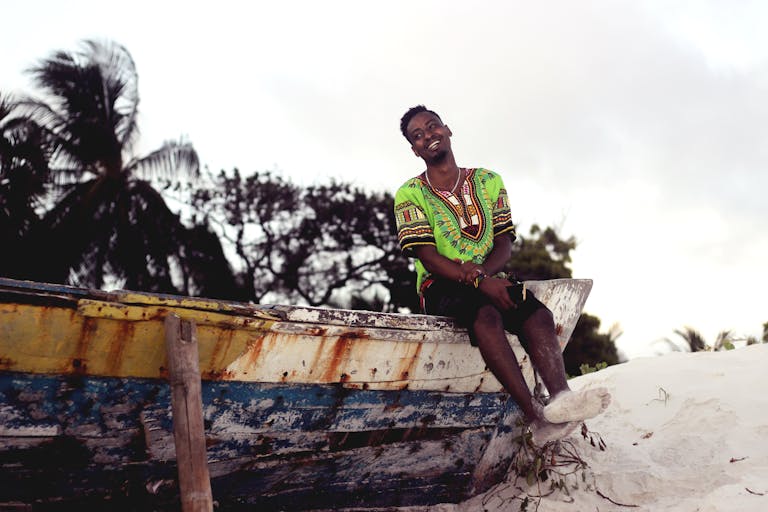 A cheerful man in colorful attire sits on a boat by the beach in Mombasa, Kenya.