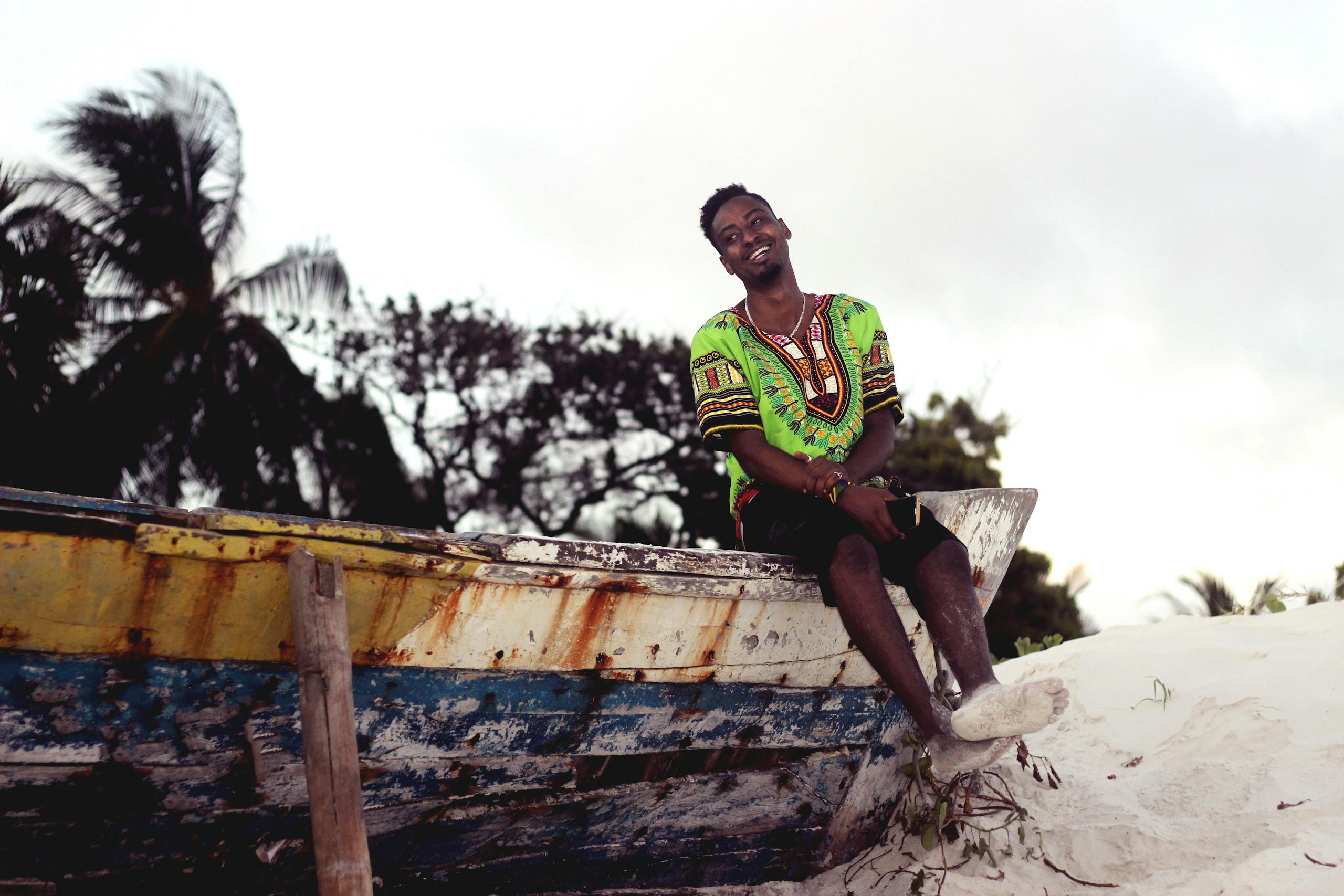 A cheerful man in colorful attire sits on a boat by the beach in Mombasa, Kenya.