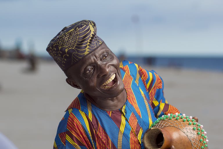 Elderly man laughing and playing a gourd shaker on a beach, wearing vibrant traditional attire.