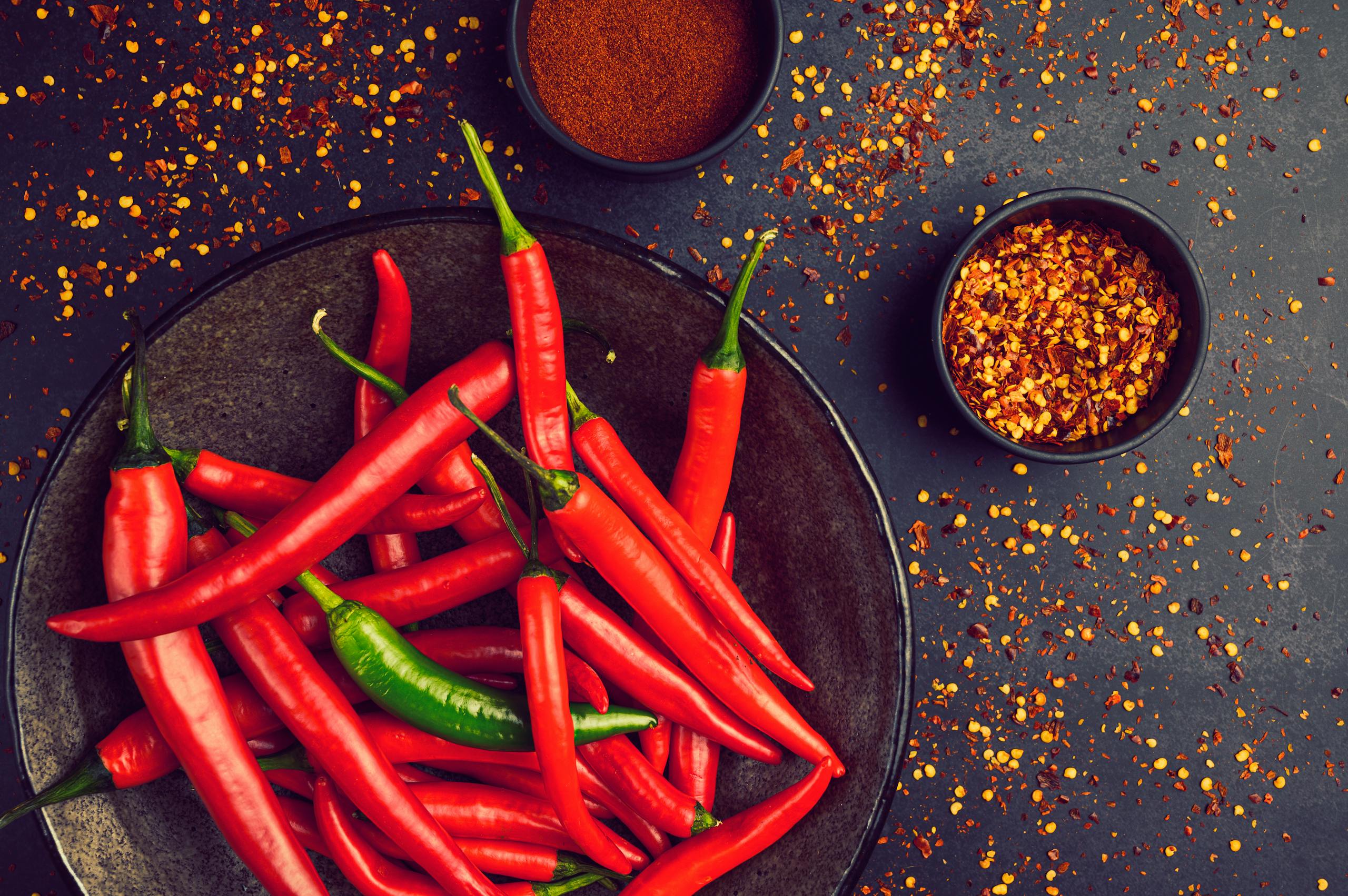 Close-up of fresh red and green chili peppers in a bowl with spices and pepper flakes.