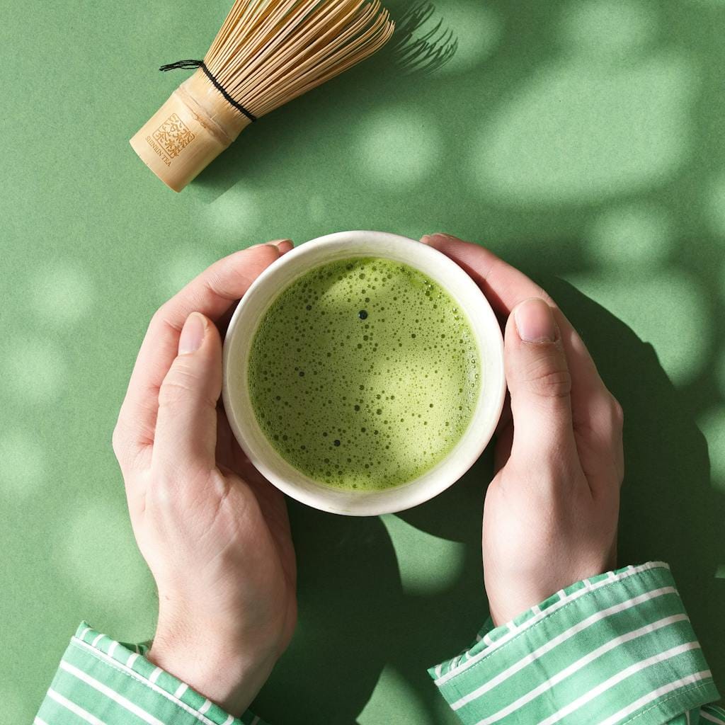 Top view of hands holding a matcha bowl and bamboo whisk on green background.