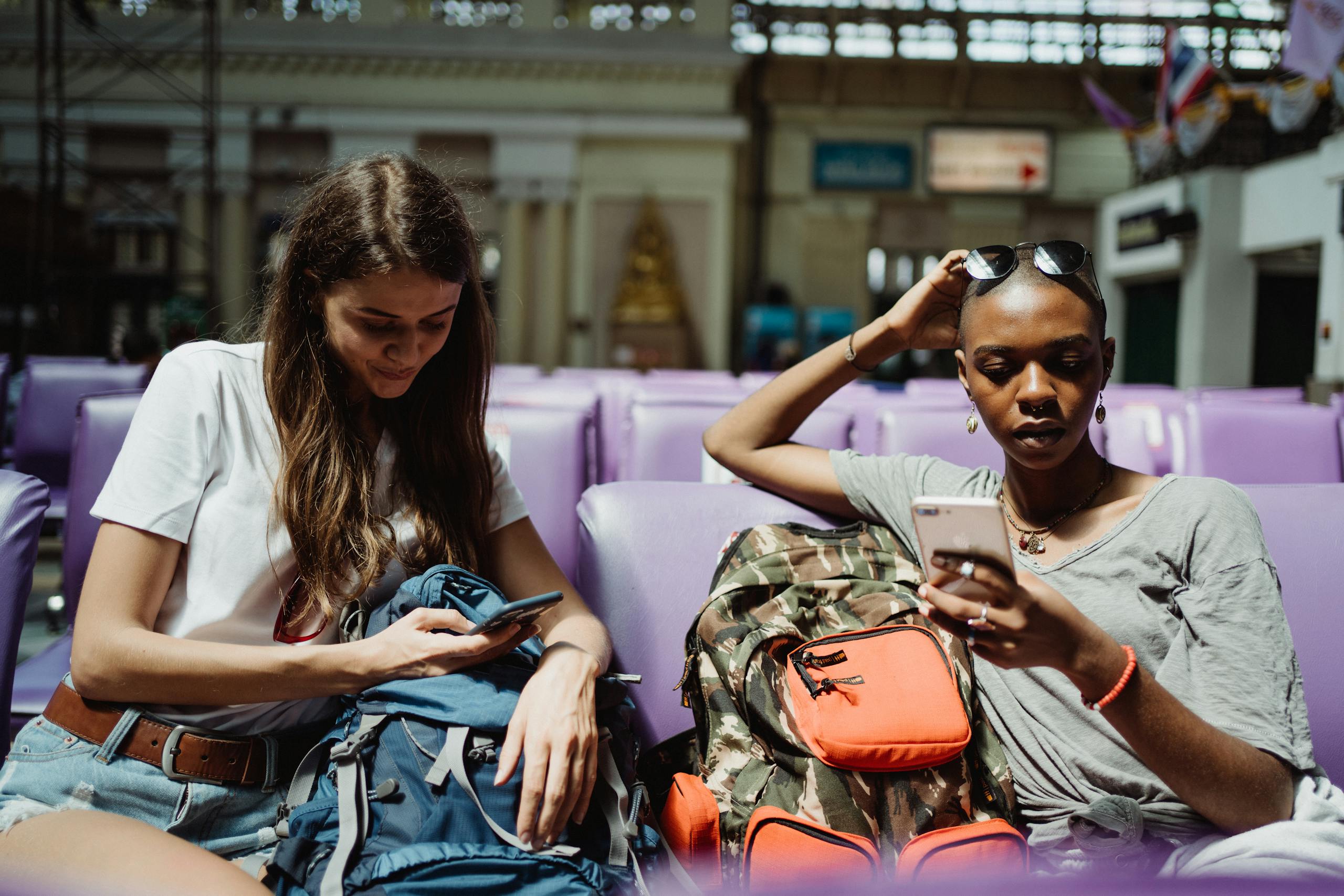 Two women with backpacks using smartphones while waiting in a railway station.